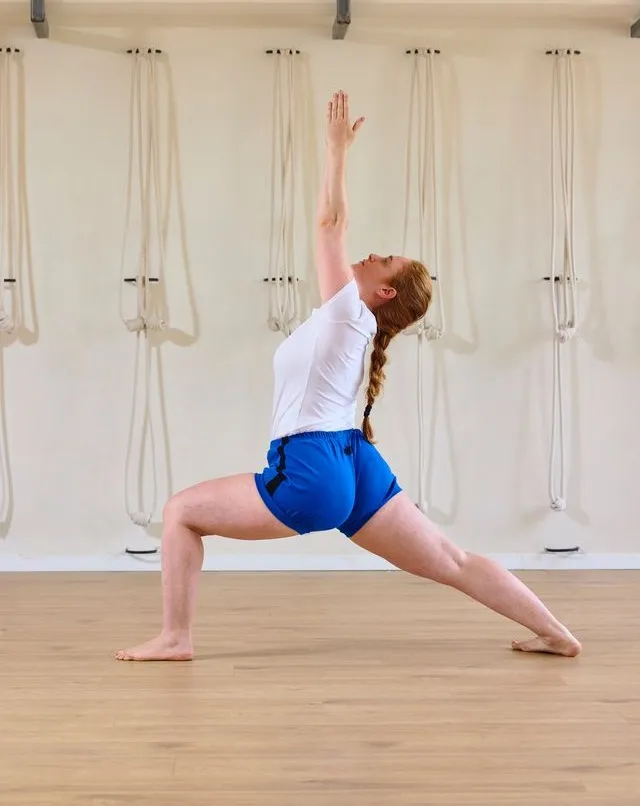 May practising Warrior I pose in the studio, arm raised overhead, beside the rope wall
