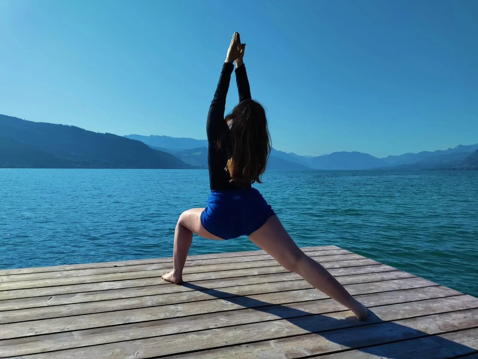 May practising Warrior I pose on a wooden dock beside an alpine lake, arms raised overhead, mountains in the background