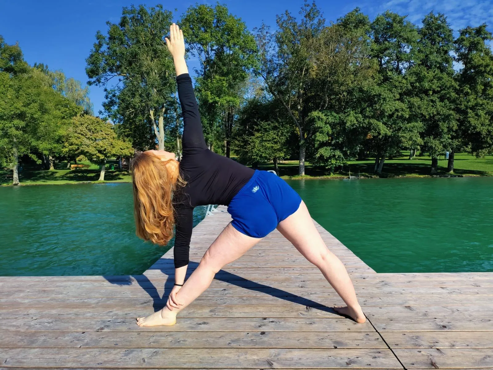 May practising Triangle pose on a wooden dock beside a calm lake, one arm reaching up